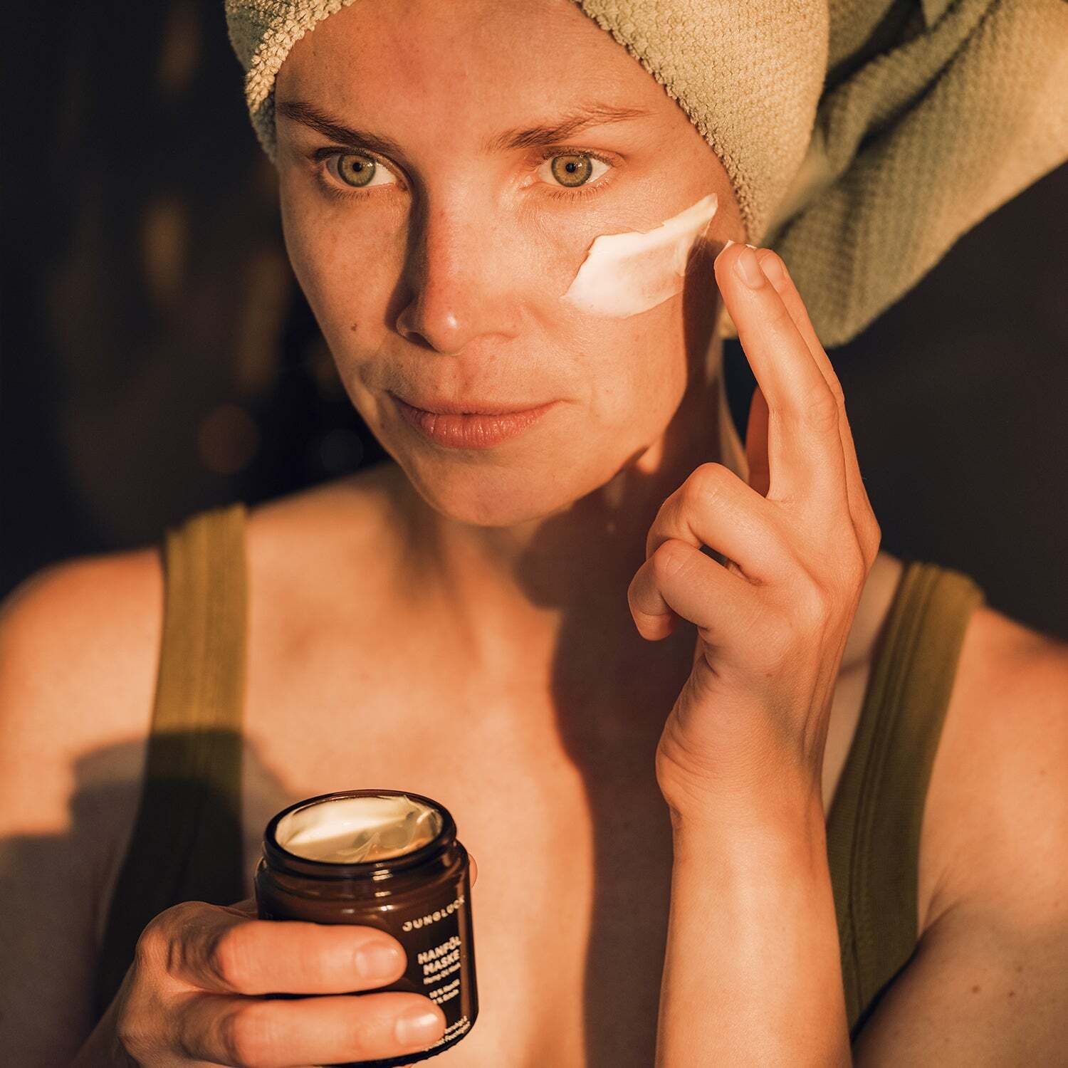 Frau mit Handtuch auf dem Kopf trägt Gesichtsmaske auf. Sie hält eine braune Dose mit der Aufschrift Hanföl Maske.