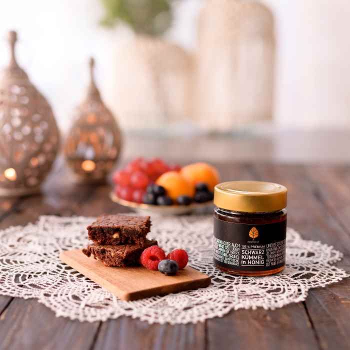 Glas mit Honig neben Brownies, Beeren und Obst. Goldener Deckel. Schwarzes Etikett mit Text: Schwarzkümmel in Honig.
