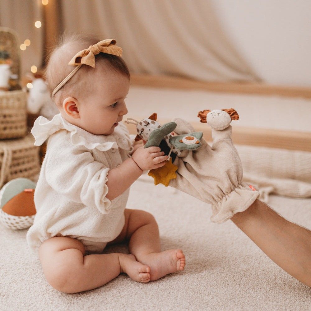 Baby spielt mit Fingerspielfigur Giraffe. Beige Stoffhandschuh mit Giraffen-, Löwen- und Blattformen. Kind sitzt auf Teppich.