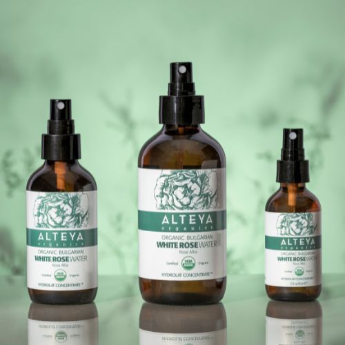 Drei braune Glasflaschen mit Sprühköpfen. Etiketten mit Text: 'ALTEYA ORGANICS White Rose Water'. Bio-Zertifizierung.