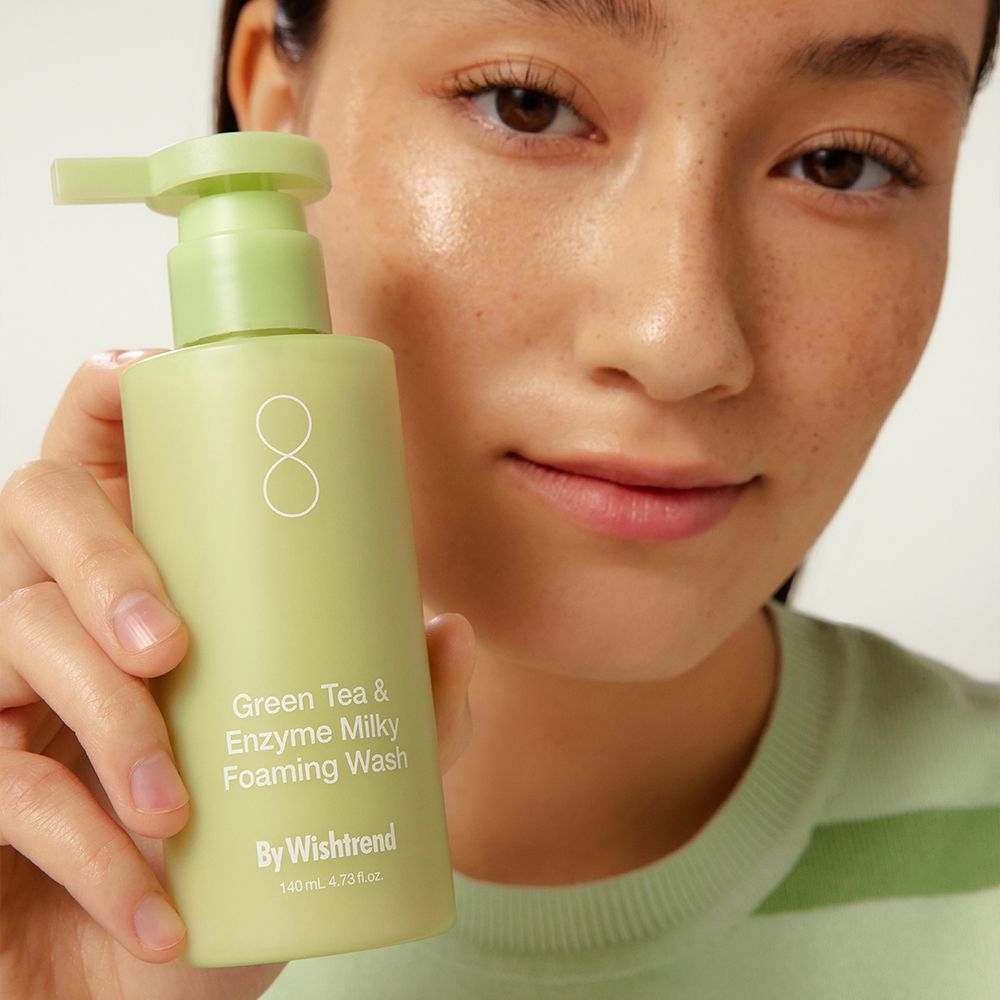 Grüne Flasche mit Pumpe. Aufschrift: Green Tea & Enzyme Milky Foaming Wash. Marke: By Wishtrend. Person hält die Flasche.