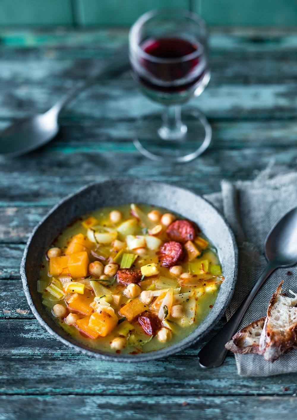 Schüssel mit Gemüsesuppe. Enthält Kichererbsen, Kürbis, Lauch, Wurstscheiben. Glas Rotwein im Hintergrund. Löffel und Brot.
