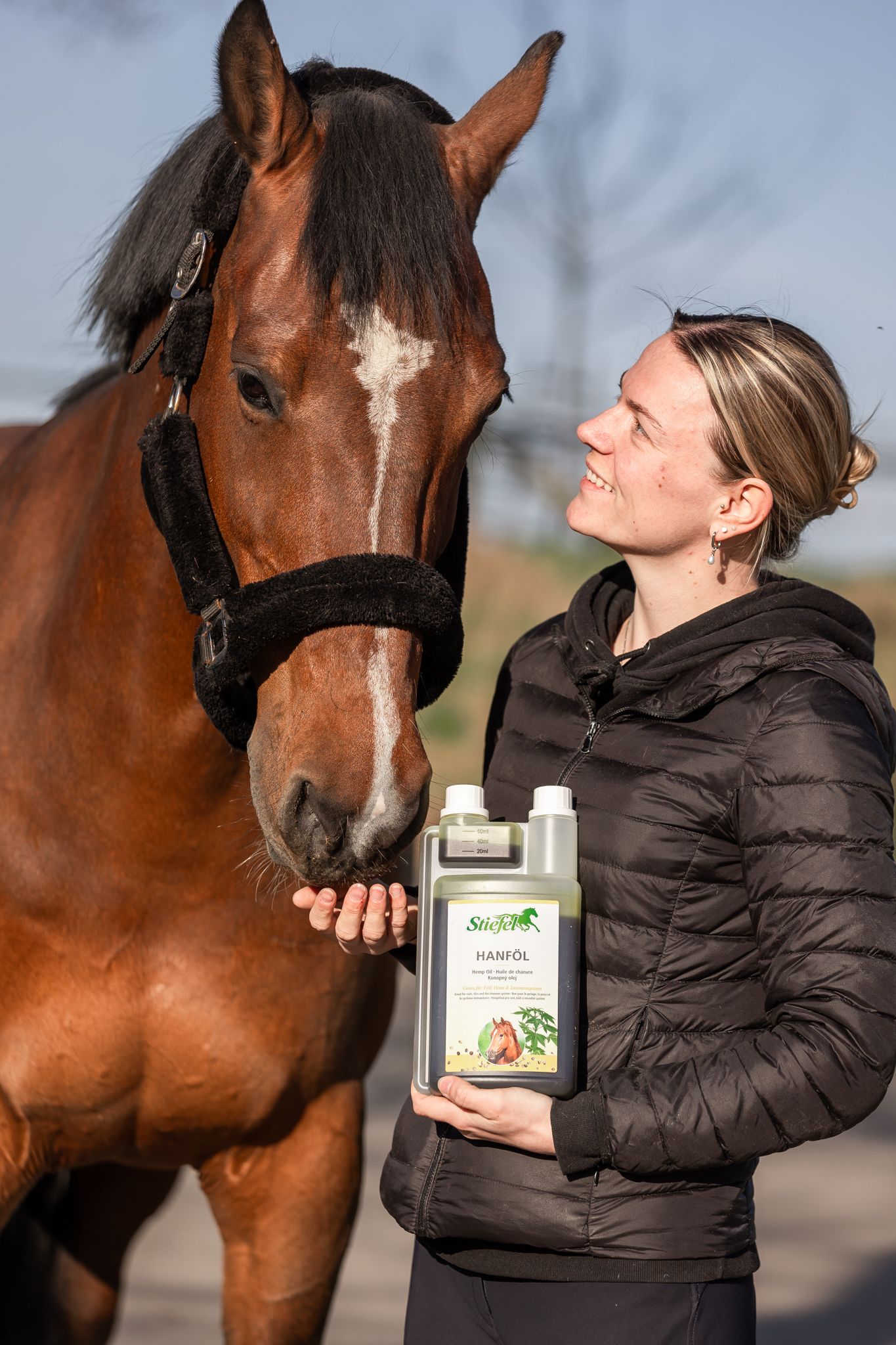 Frau und Pferd stehen nebeneinander. Die Frau hält eine Flasche Stiefel Hanföl. Das Pferd schaut zur Frau.