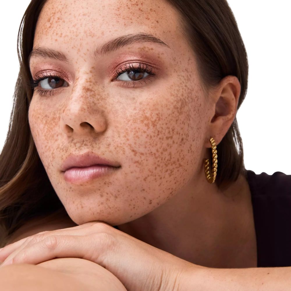 Porträt einer Frau mit Sommersprossen. Sie trägt rosafarbenen Lidschatten und goldene Creolen.