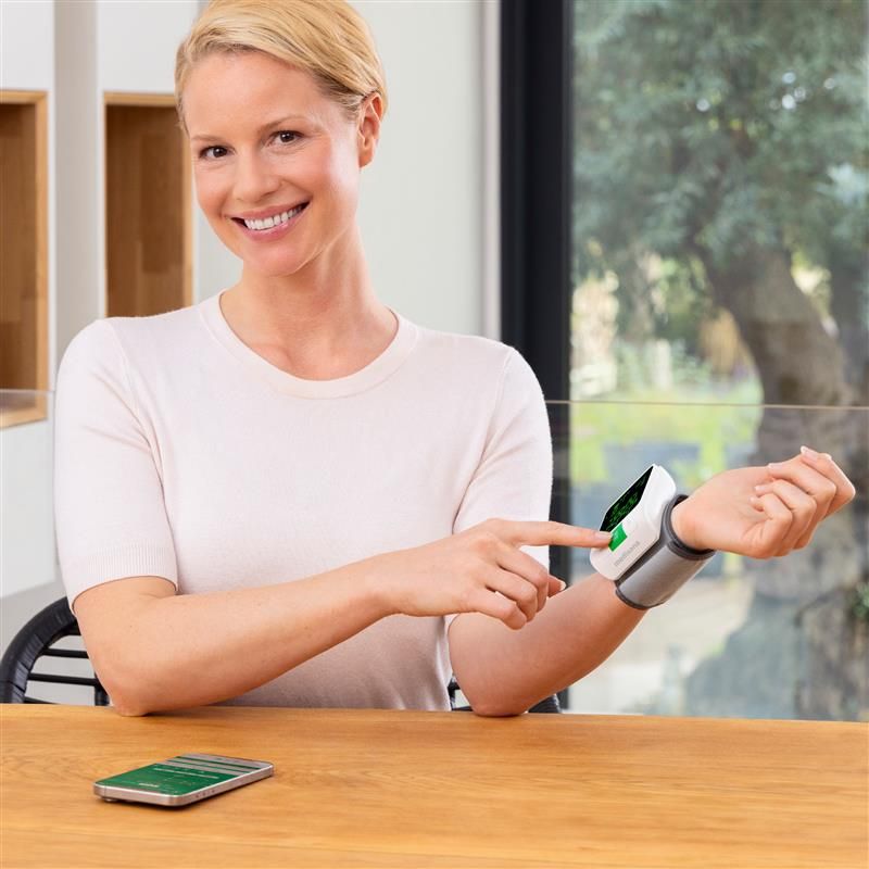 Frau mit Handgelenk-Blutdruckmessgerät. Zeigt auf Gerät. Smartphone auf Tisch. Helles Holz. Fenster im Hintergrund.