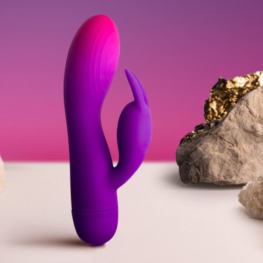 Lila Vibrator mit rosa Spitze und Hasenohren. Lila-rosa Farbverlauf. Auf weißem Untergrund mit Steinen im Hintergrund.
