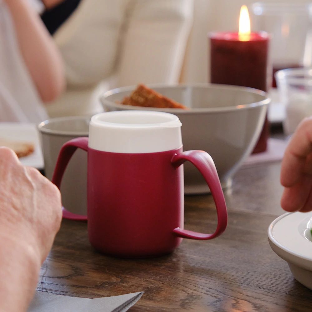 Becher mit zwei Henkeln, rot, weißer Rand. Auf einem Tisch, im Hintergrund Essen.