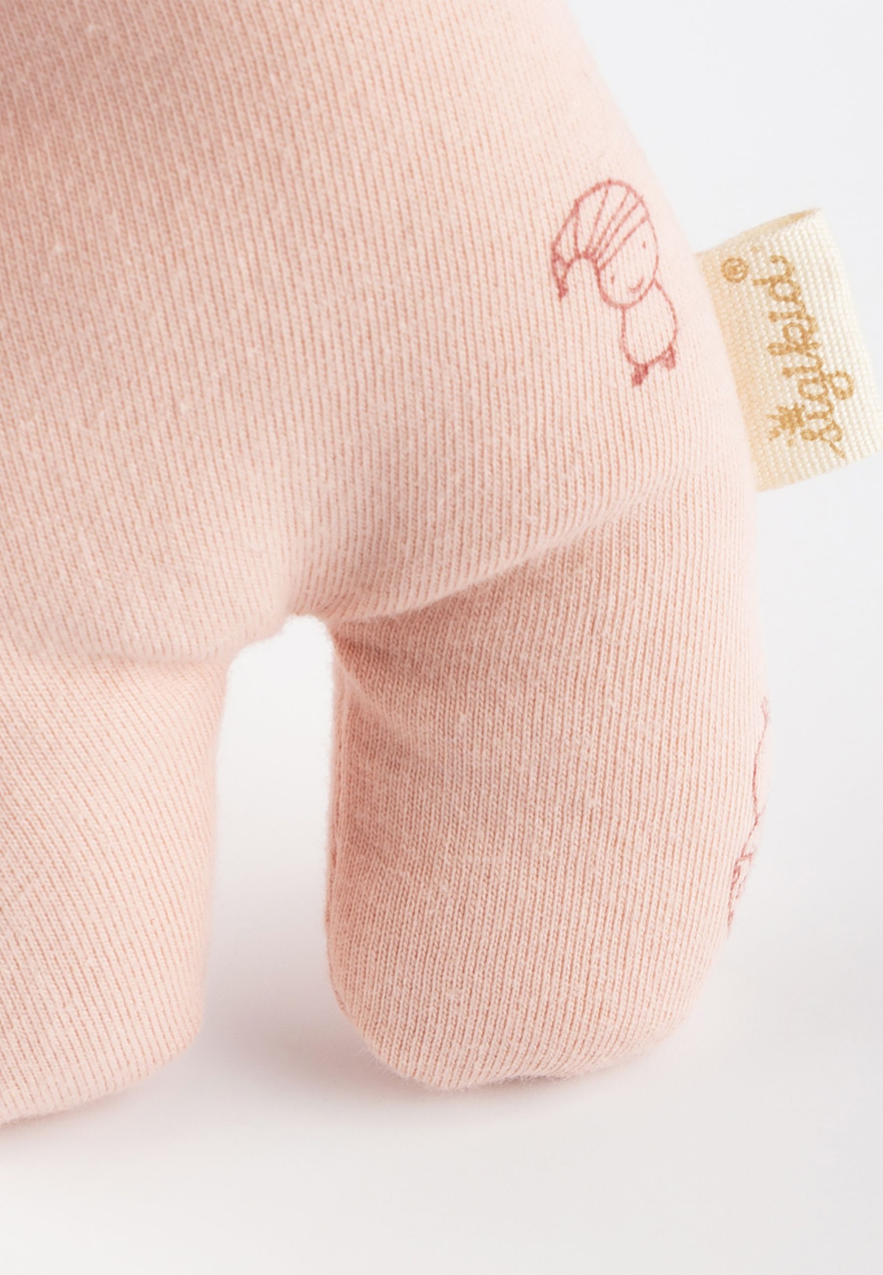 Detail der Puppe. Rosa Stoff, Etikett mit Logo. Aufgenähte Illustration. Naht am Bein sichtbar.
