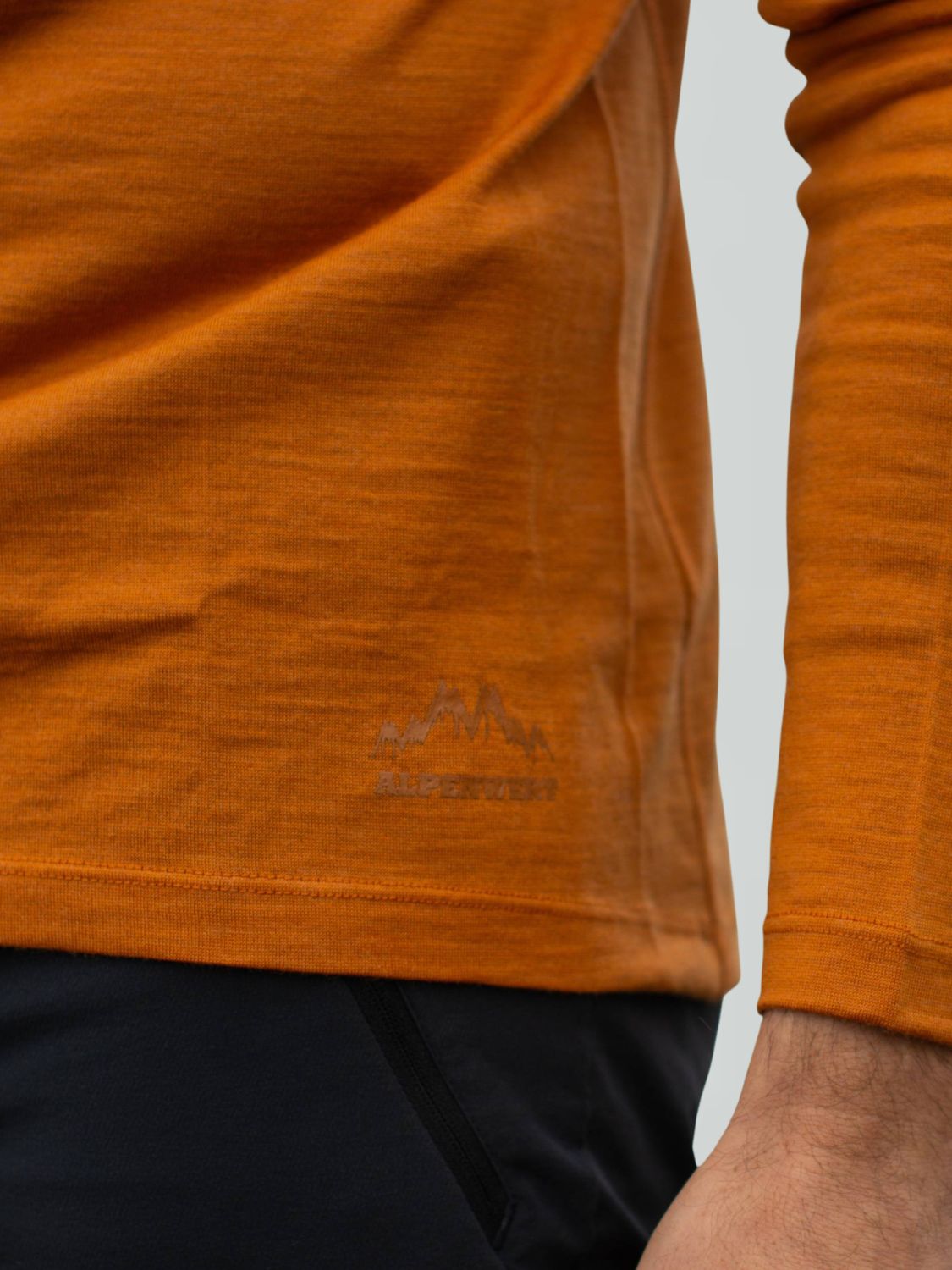 Detailansicht des orangefarbenen Merino-Pullovers mit kleinem Berg-Logo am Saum.