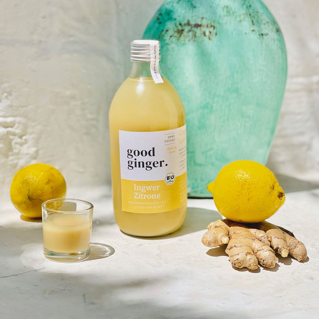 Glasflasche mit gelbem Inhalt, daneben ein Shot-Glas, Zitronen und Ingwer. Etikett "good ginger.", "Ingwer Zitrone" und Bio-Siegel.