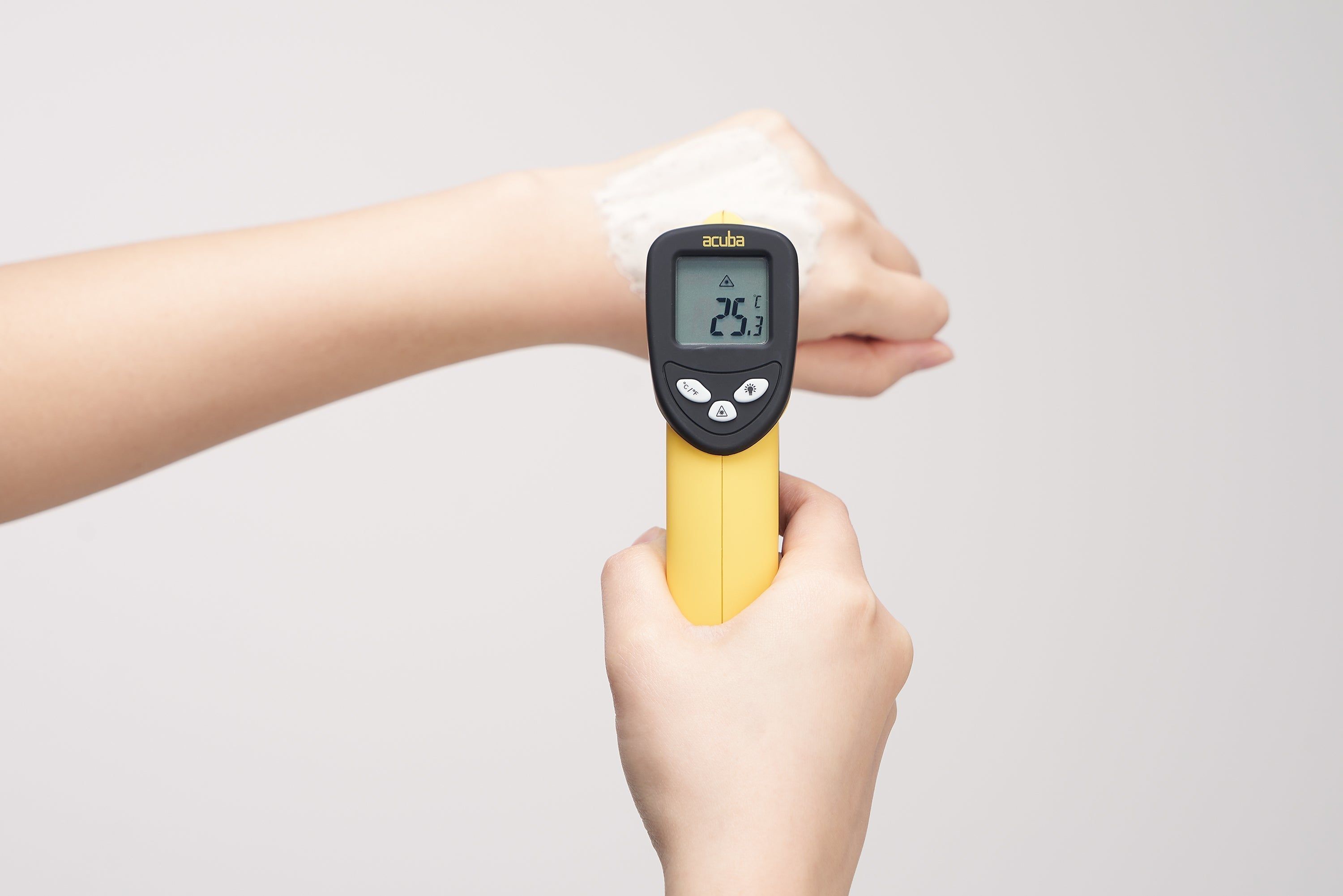 Hand hält ein gelbes Thermometer an einem Arm. Anzeige: 25.1. Auf weißem Hintergrund.
