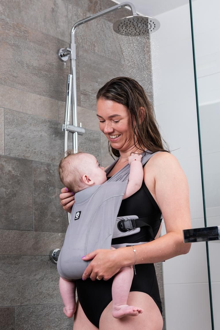 Frau mit Baby in grauer Babytrage unter der Dusche. Dooky-Logo sichtbar.