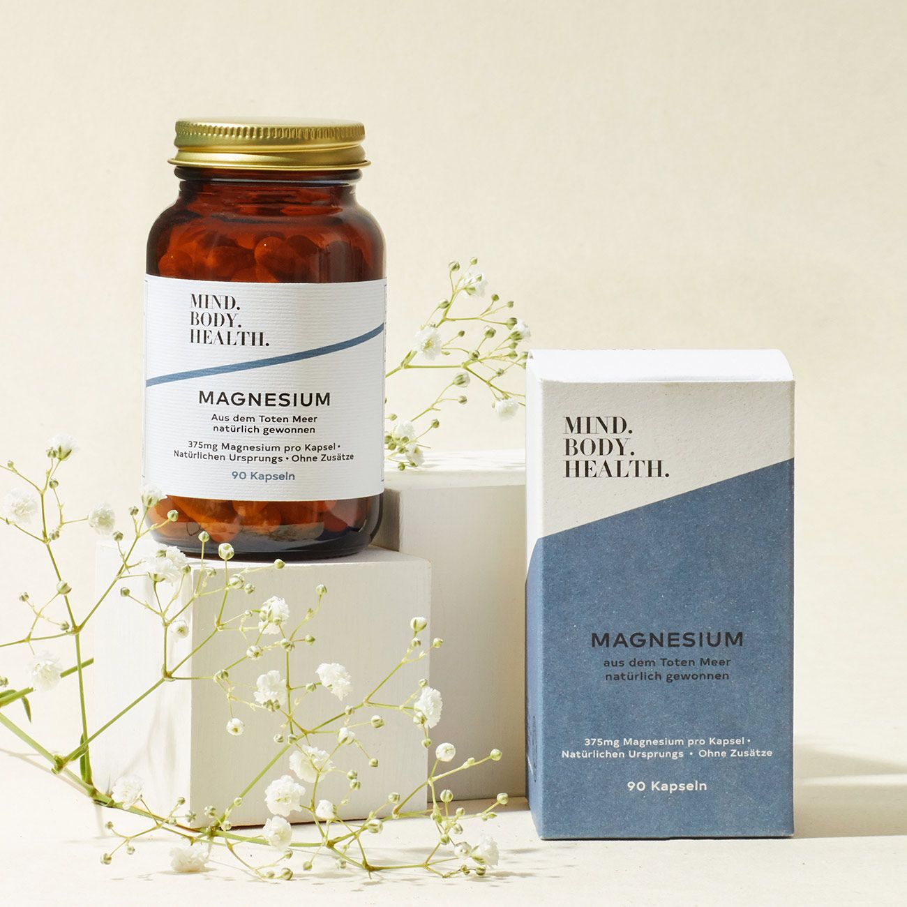 Produktpräsentation mit Flasche und Schachtel. Flasche mit "MIND.BODY. HEALTH" und "Magnesium". Schachtel mit gleichem Design. Weiße Blüten.