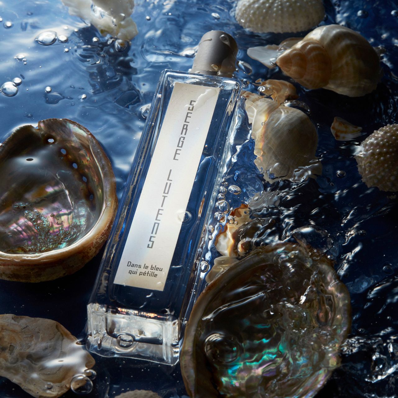 Rechteckige Glasflasche im Wasser, umgeben von Muscheln. Aufschrift SERGE LUTENS. Unterhalb: Dans le bleu qui pétille.