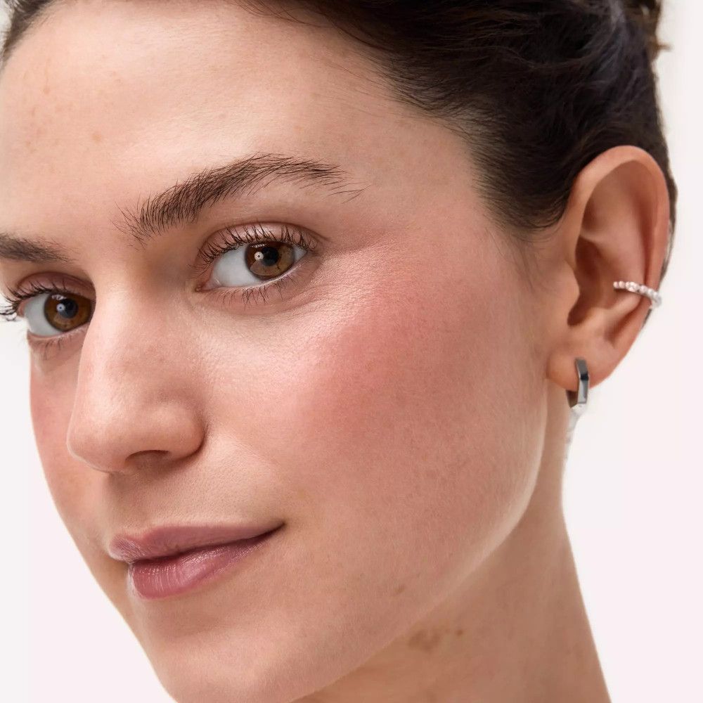 Nahaufnahme eines Gesichts mit Rouge auf den Wangen. Die Person trägt Ohrringe.