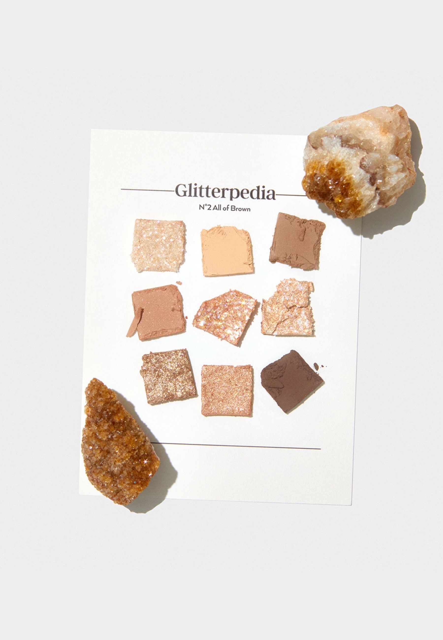 Glitterpedia N°2 All of Brown Lidschatten. Neun Farbmuster auf weißem Papier. Zwei Steine liegen daneben.