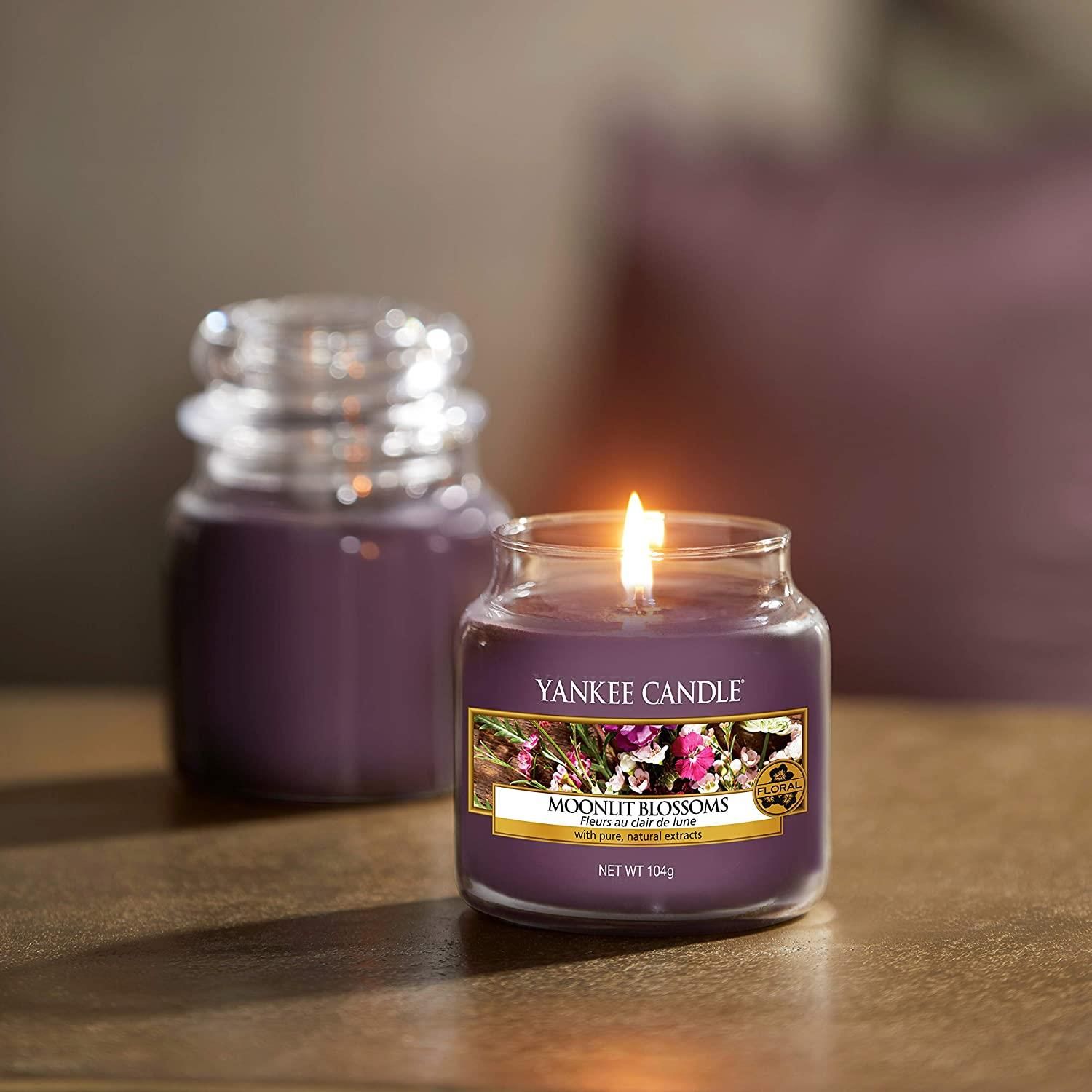Zwei Duftkerzen auf Holzoberfläche. Eine brennt, Etikett sichtbar. Violettes Wachs, Glasbehälter.