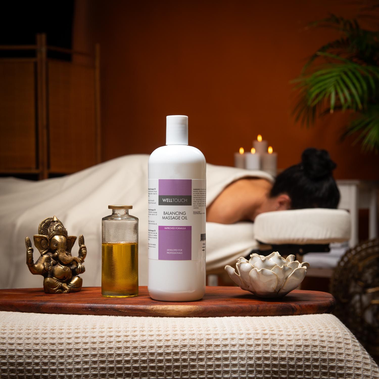 Weißes Welltouch Massageöl, daneben eine kleine Glasflasche mit Öl. Im Hintergrund eine Person auf einer Massageliege.