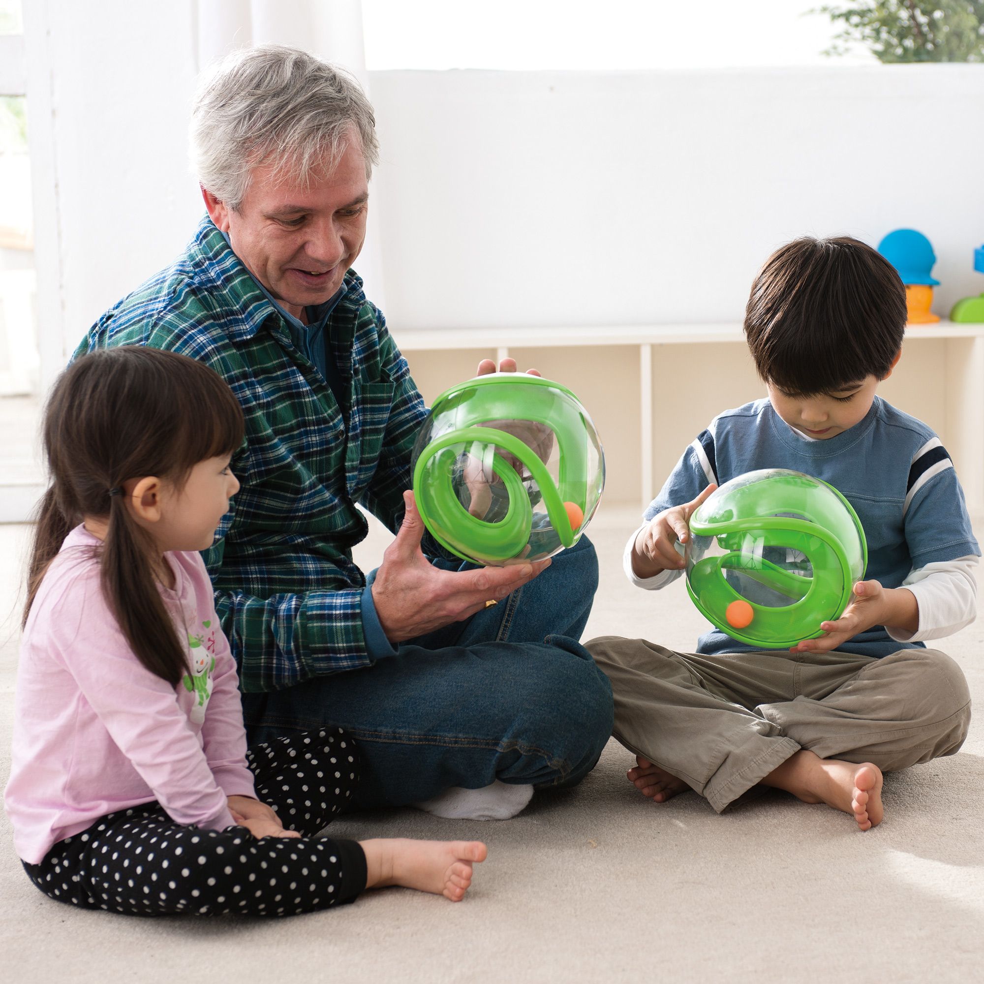 Drei Personen halten grüne, transparente Bälle. Erwachsene und Kinder sitzen auf dem Boden. Helle Umgebung.