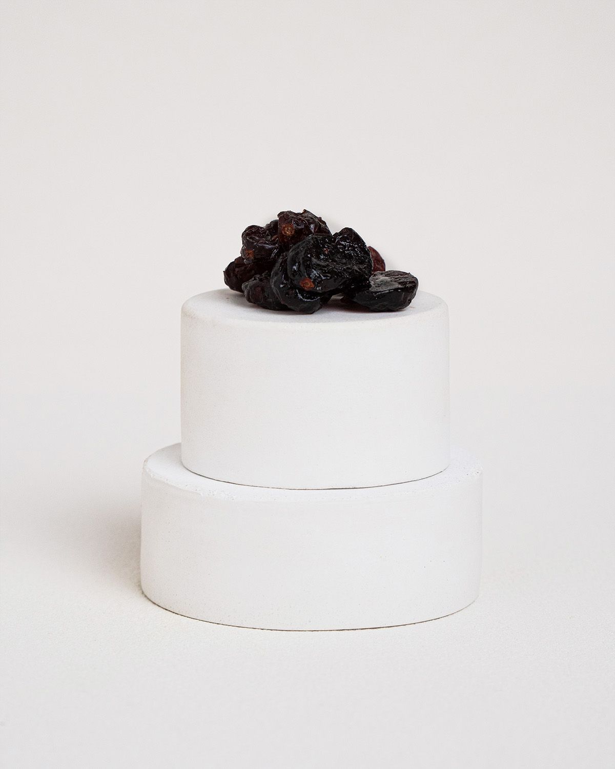 Getrocknete Cranberries auf weißem, zylindrischen Podest. Dunkelrote Farbe, unregelmäßige Form.