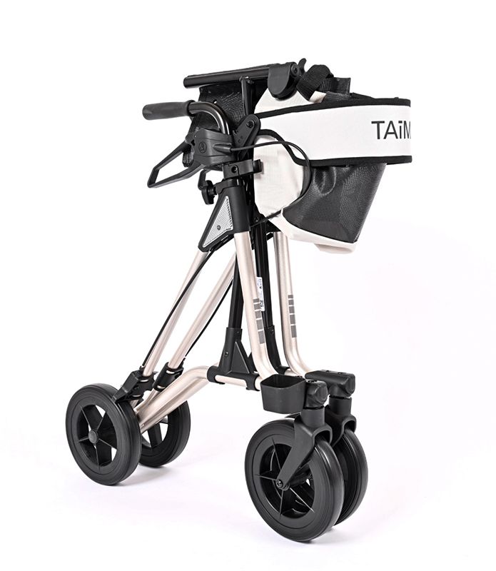 Zusammengeklappter Rollator mit vier Rädern. Der Rollator hat einen schwarzen Korb und die Sitzfläche ist weiß. Der Schriftzug TAiMA ist sichtbar.