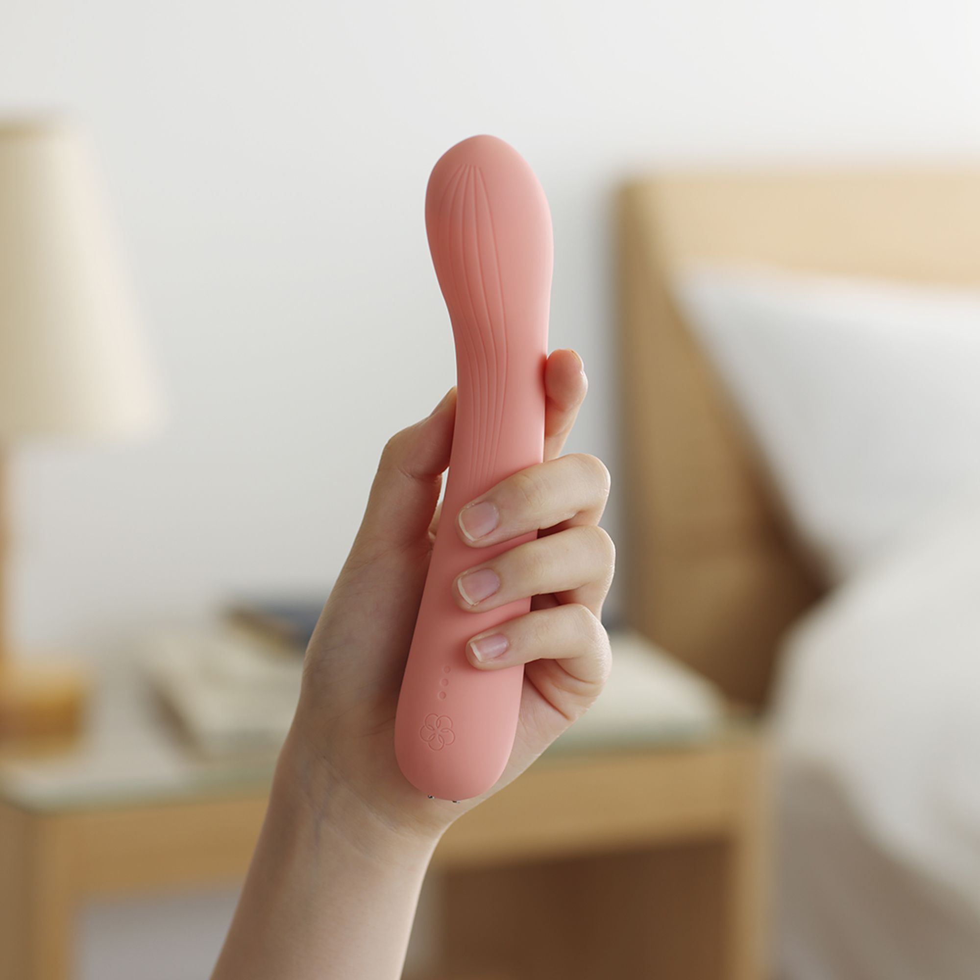 Hand hält rosa Vibrator. Produktname nicht sichtbar. Schlafzimmer-Hintergrund.