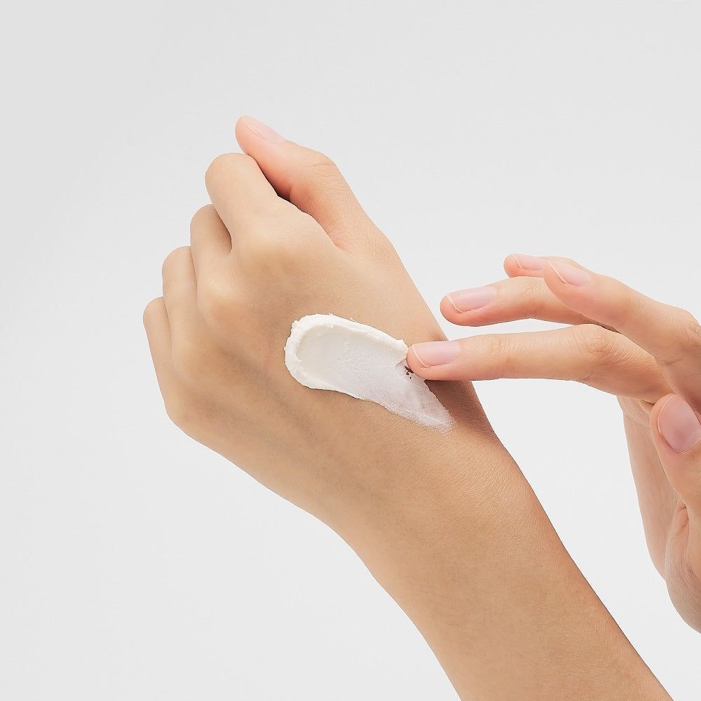 Eine Hand mit einer weißen Creme-Probe auf dem Handrücken, die mit einem Finger verrieben wird.