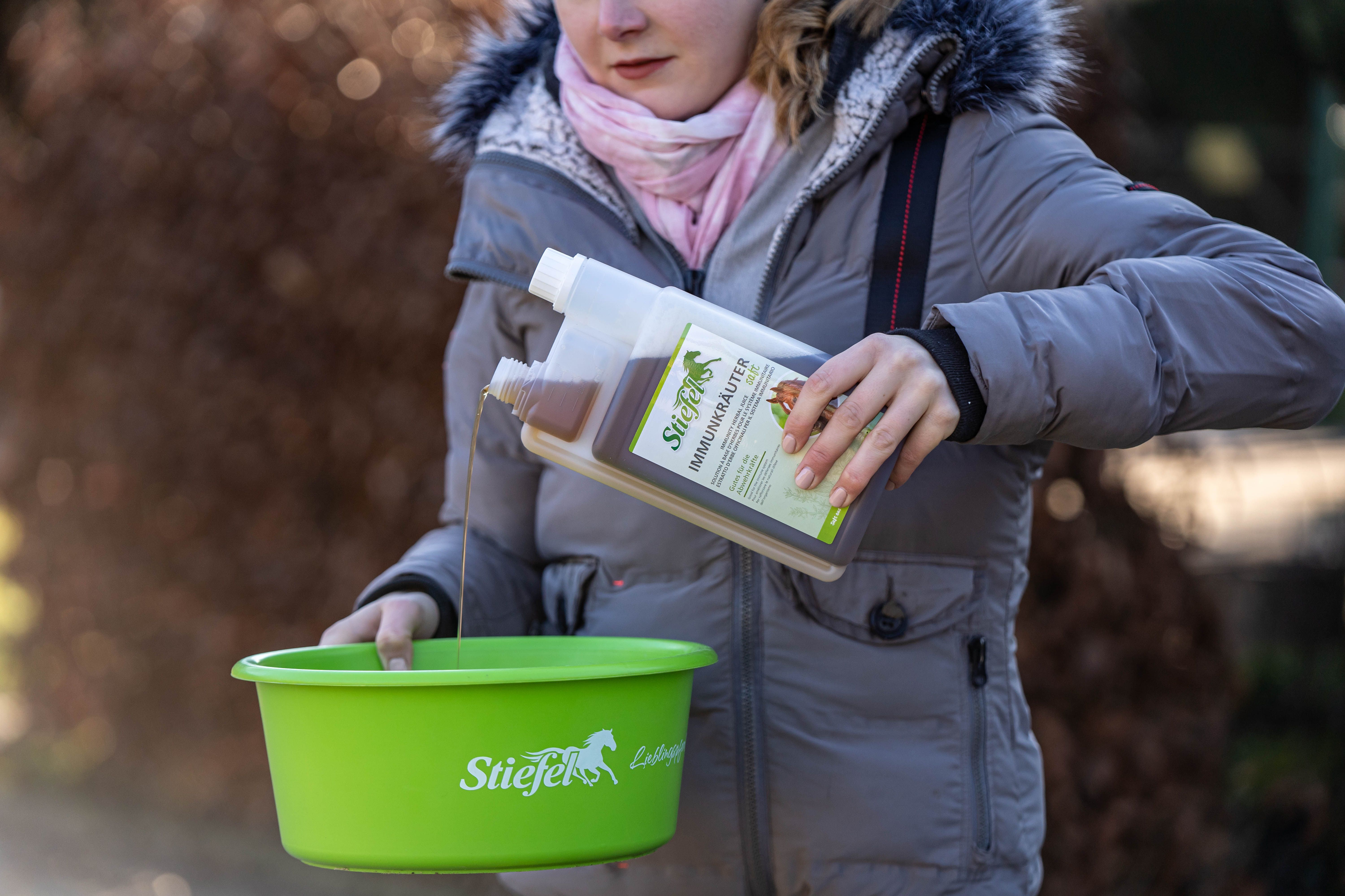 Person hält eine Flasche mit brauner Flüssigkeit und eine grüne Schüssel. Flasche mit weißem Deckel und Etikett. Schüssel mit Logo.