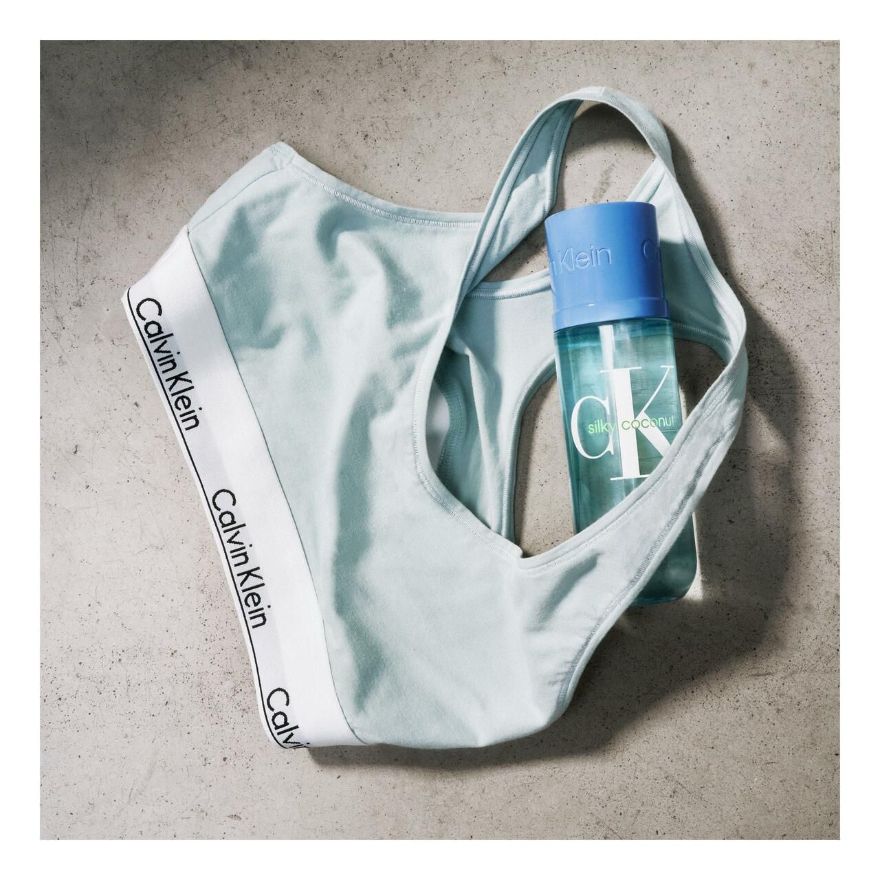 Hellblaues Sport-BH mit weißem Calvin Klein-Logo. Daneben eine hellblaue Flasche mit blauem Deckel. Aufschrift "Calvin Klein".
