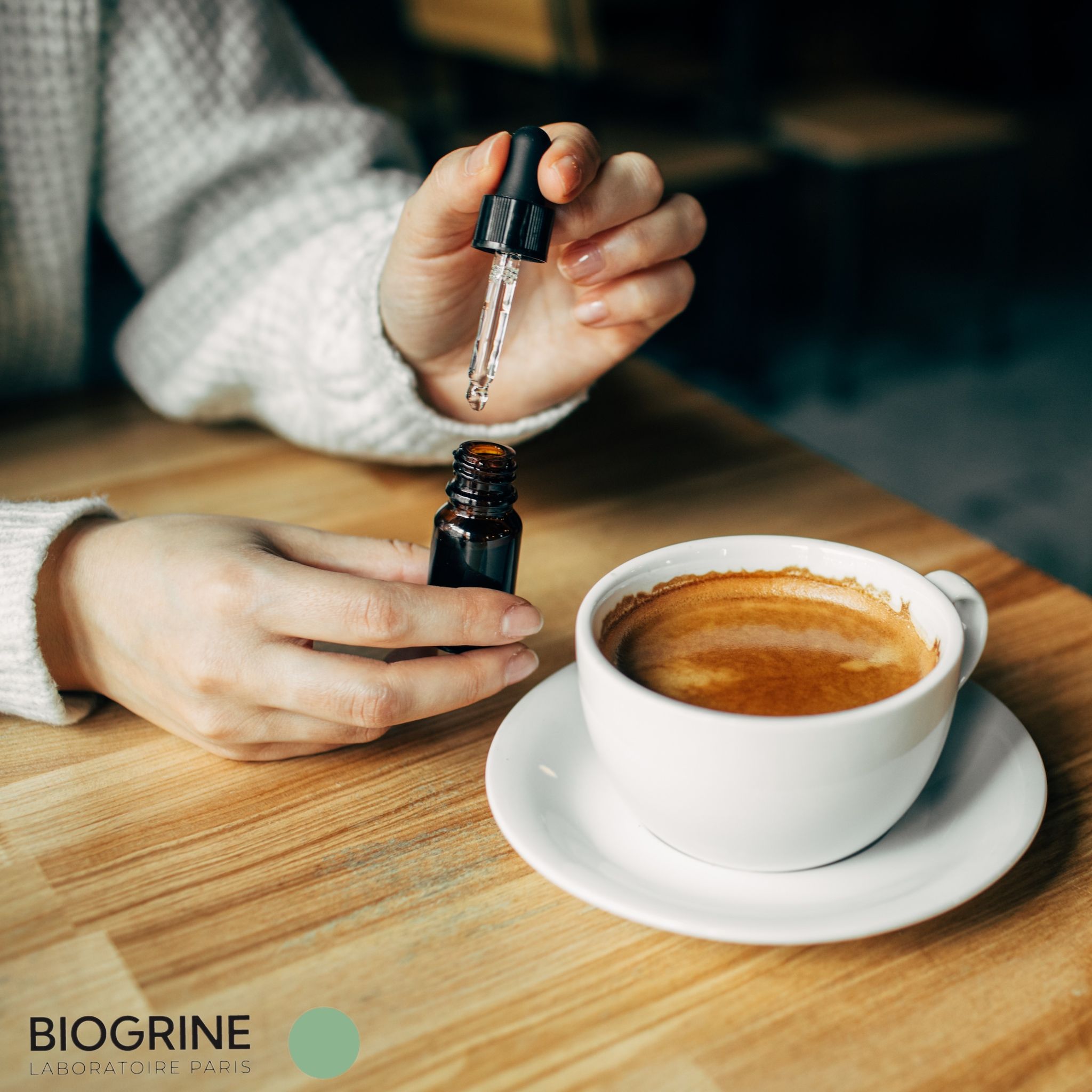 Hände halten eine Pipette über einer kleinen braunen Flasche und einer Kaffeetasse auf einem Tisch.