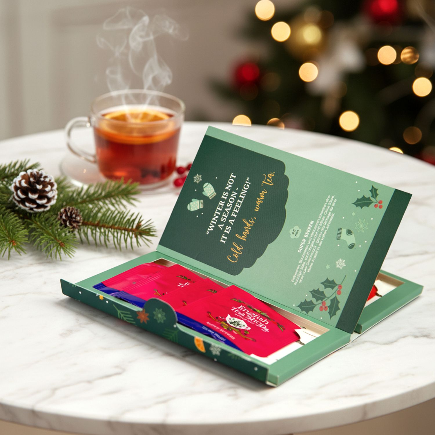 Geöffnete grüne Geschenkbox mit Teebeuteln. Aufschrift: English Tea Shop. Zitat: Winter ist keine Jahreszeit, es ist ein Gefühl. Tasse Tee daneben.