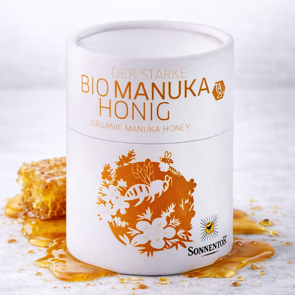 Bio Manuka Honig in weißer Dose mit Bienenmotiv. Honig tropft von einem Holzlöffel auf Brotscheiben.