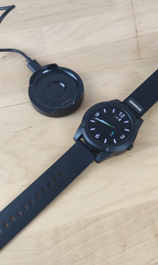 Schwarze Smartwatch mit Silikonarmband und Ladekabel auf Holzhintergrund.