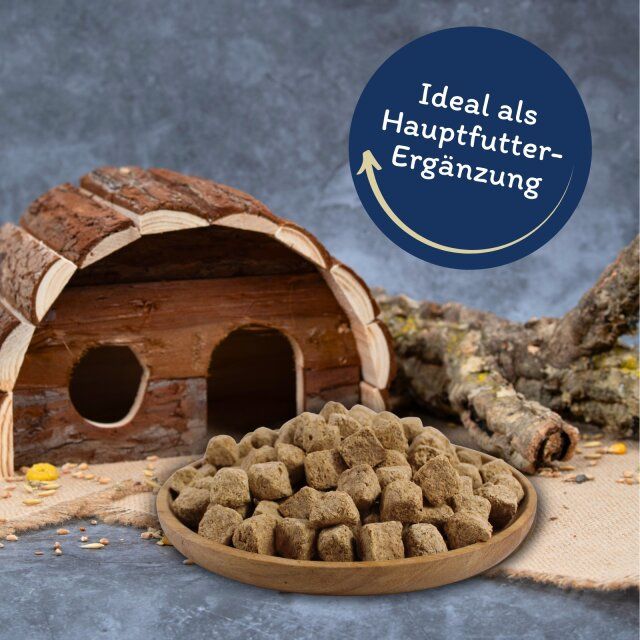 Gefriergetrocknete Würfel in einer Holzschale. Beschriftung: Ideal als Hauptfutter-Ergänzung. Hintergrund: Haus.