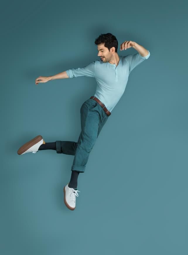 Mann in hellblauem Hemd und Jeans, springt. Er trägt schwarze Socken und weiße Schuhe. Blauer Hintergrund.