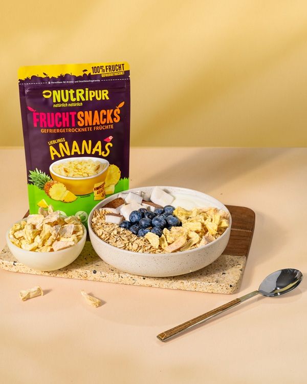 Produktpräsentation mit gefriergetrockneten Ananas-Bits, Blaubeeren, Kokosraspeln und Müsli in Schalen. Verpackung im Hintergrund.