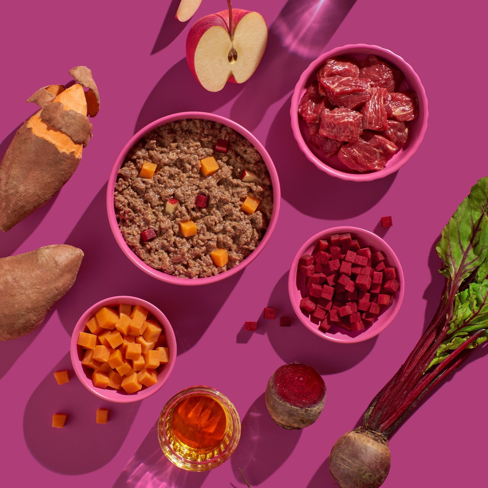 Verschiedene Zutaten in Schalen: Fleisch, Süßkartoffeln, Rote Bete, Apfelstücke. Rosa Hintergrund.