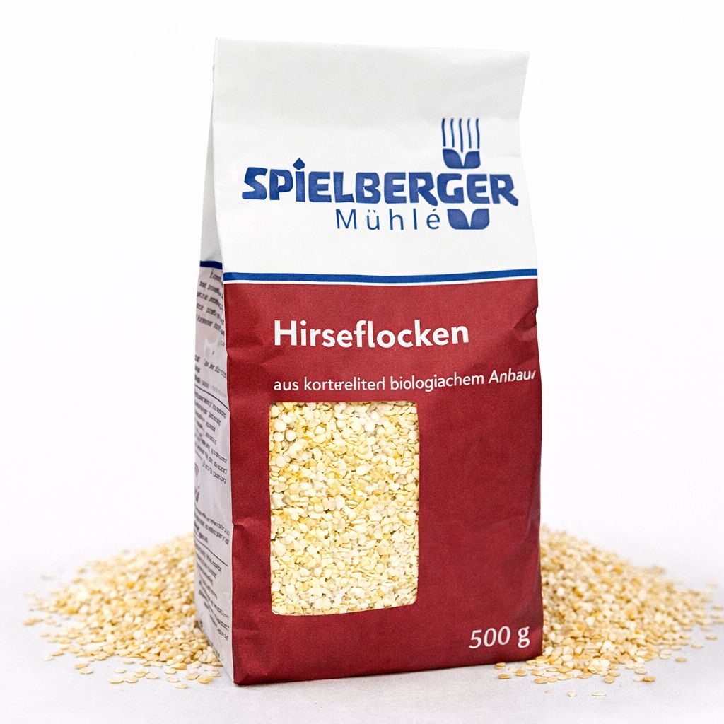Packung Spielberger Mühle Hirseflocken (500g) mit Sichtfenster, umgeben von verstreuten Hirseflocken.
