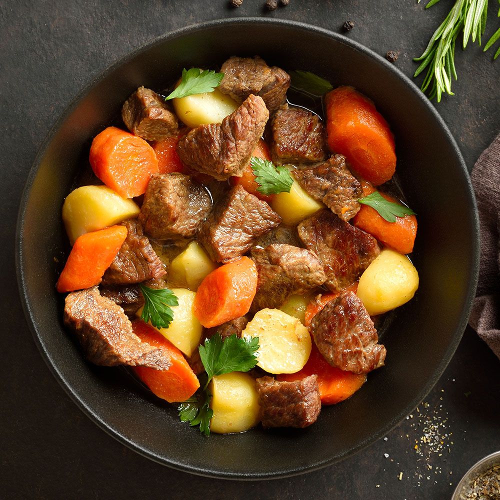 Eintopf mit Fleisch, Karotten und Kartoffeln in schwarzer Schüssel. Petersilie als Garnitur.