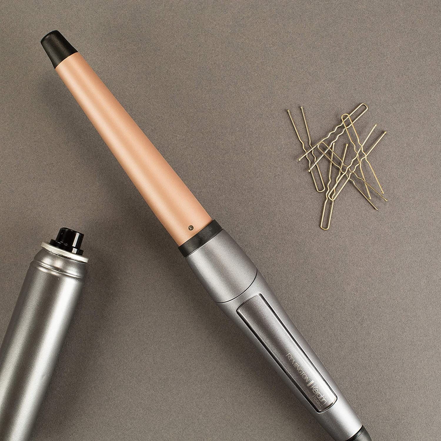 Lockenstab mit silbernem Griff und roségoldfarbener Spitze. Daneben eine Haarspraydose und Haarnadeln.