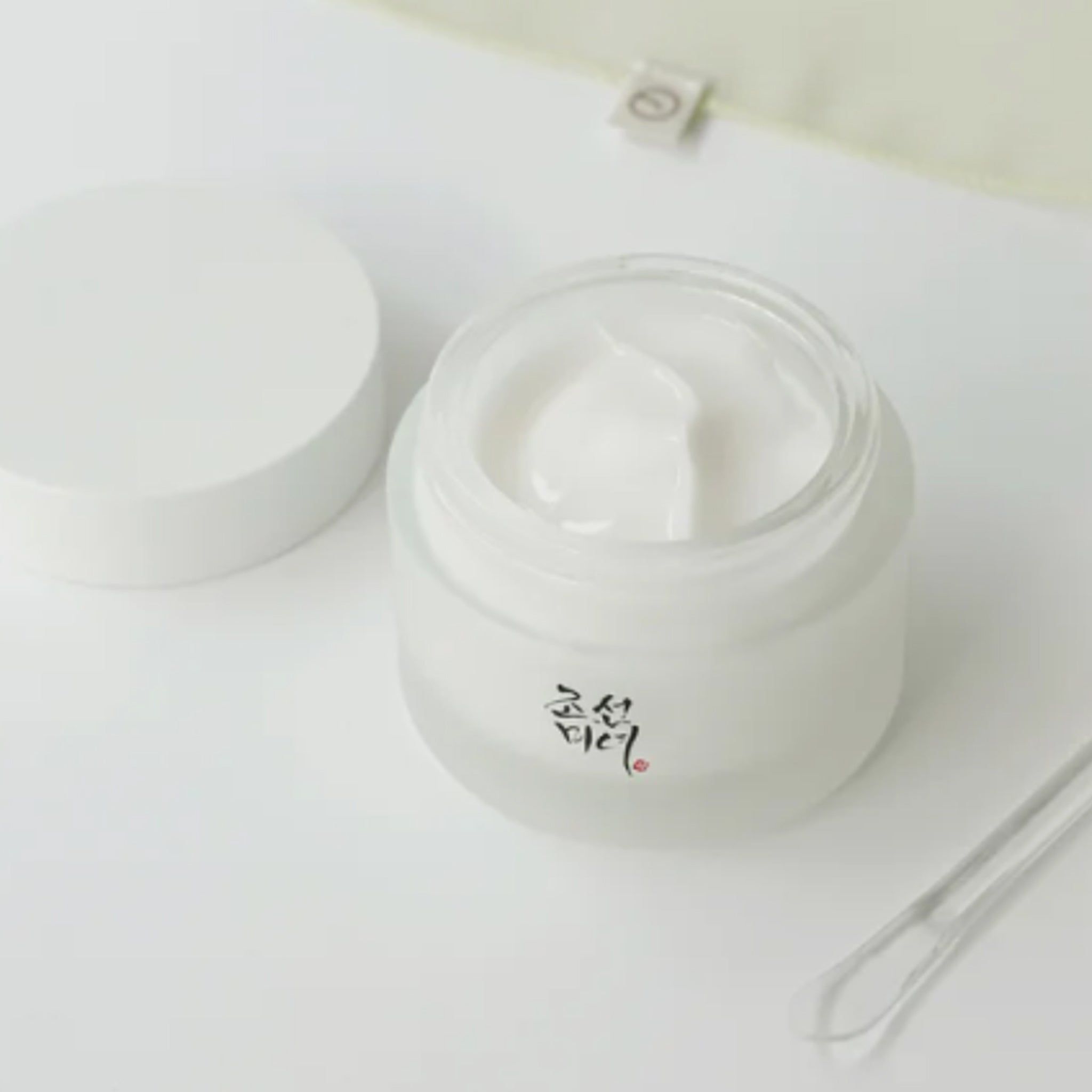 Glas mit Creme, Deckel daneben. Aufschrift in koreanischer Schrift. Spatel liegt daneben.