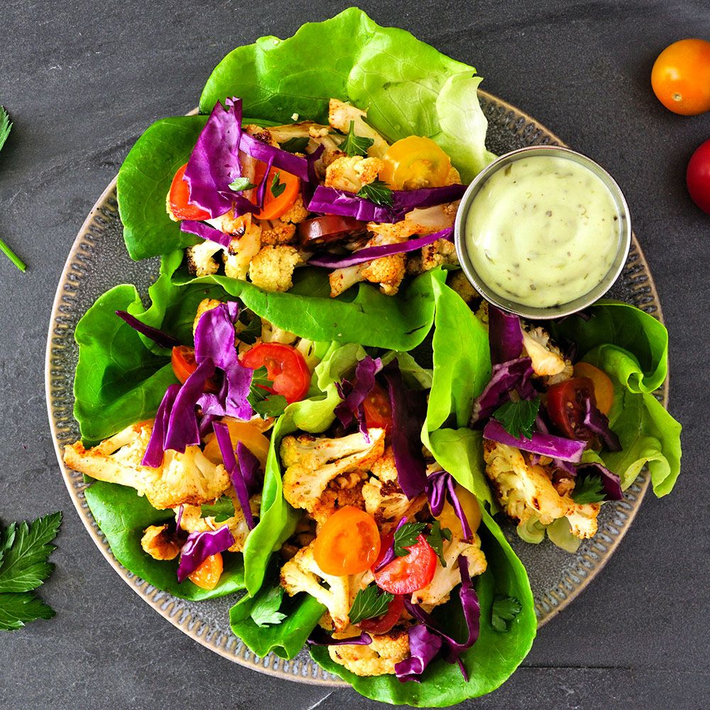 Gemüsetacos mit Blumenkohl, Rotkohl, Tomaten und Sauce auf einem Teller. Salatblätter als Hülle. Grauer Hintergrund.