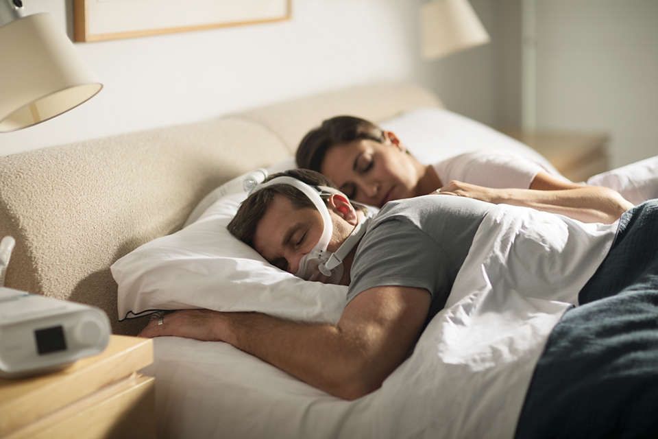 Mann schläft im Bett mit CPAP-Maske, Frau schläft daneben. CPAP-Gerät auf Nachttisch.