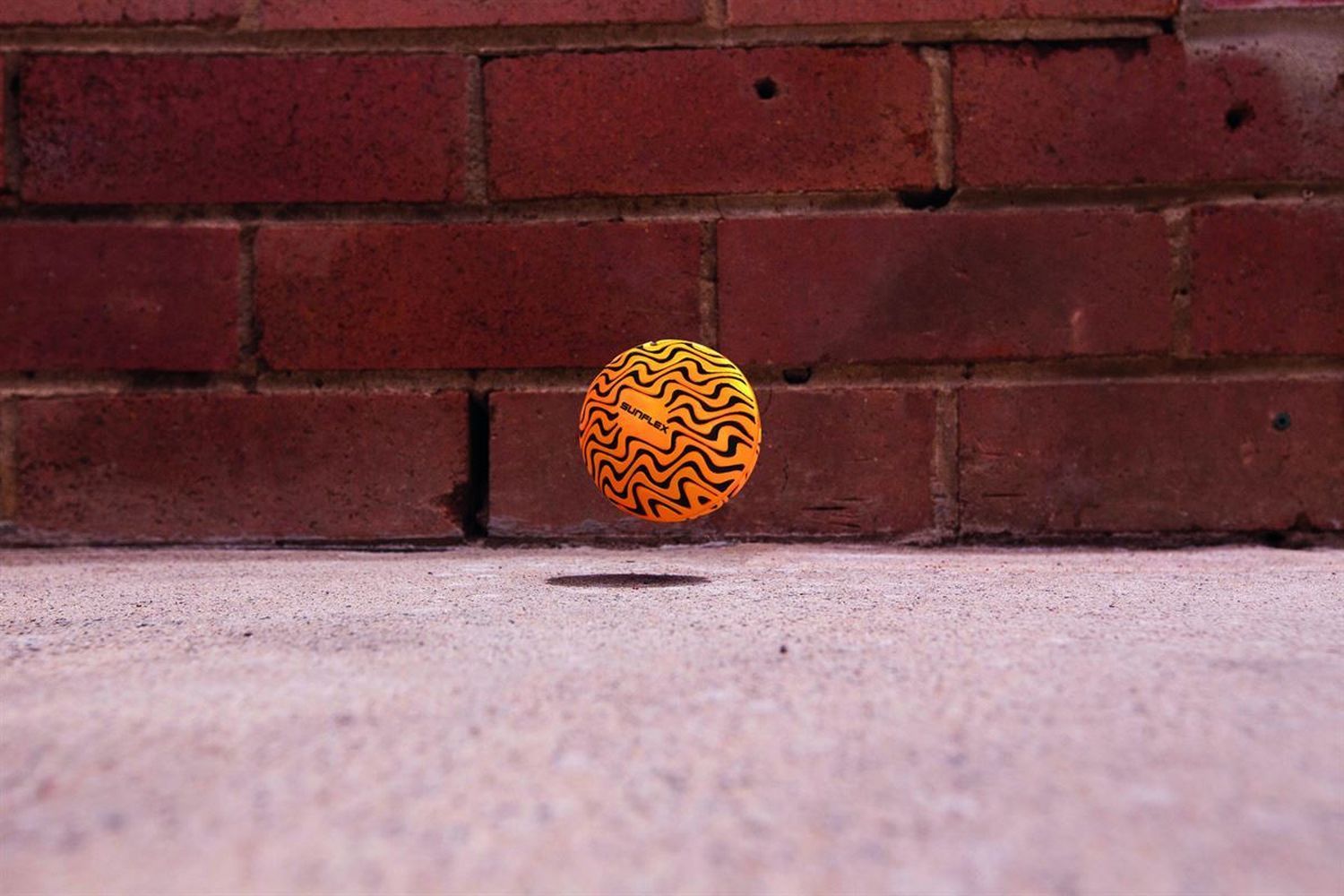 Schwebender orangefarbener Ball mit schwarzen wellenförmigen Linien vor einer Backsteinmauer.