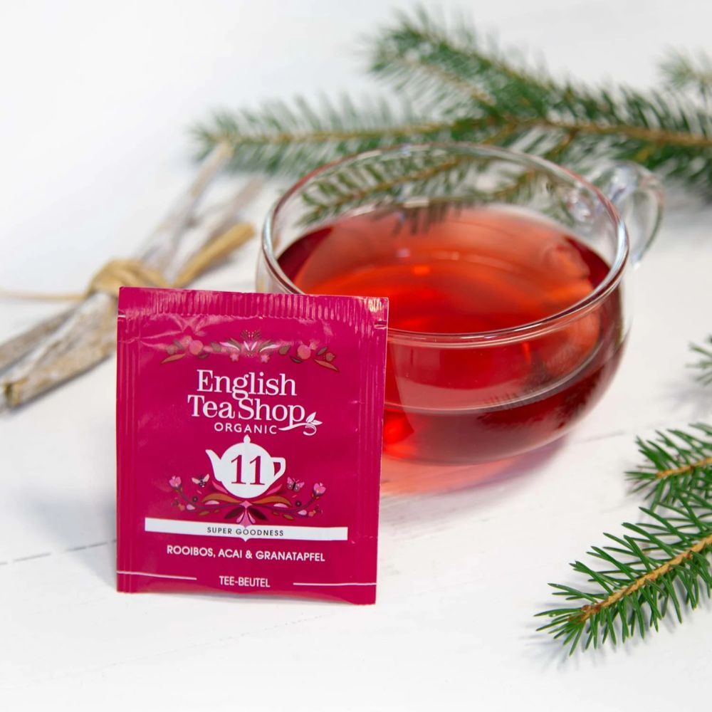 Teebeutel mit Aufdruck "11 Rooibos, Acai & Granatapfel". Marke: English Tea Shop. Tee in Glas. Glas mit rotem Tee. Grünzweig daneben.