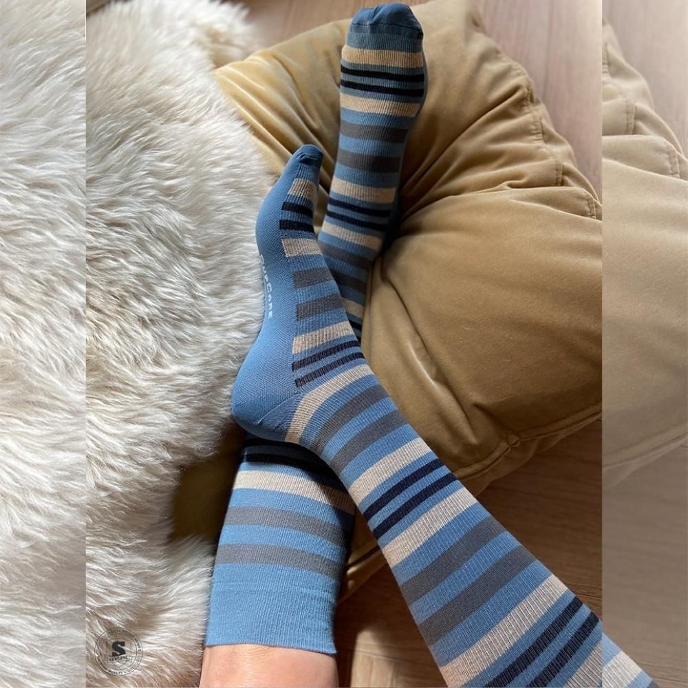 Person trägt blau-beige gestreifte Stützstrümpfe. Die Strümpfe sind auf einem Kissen und einem Fell platziert.