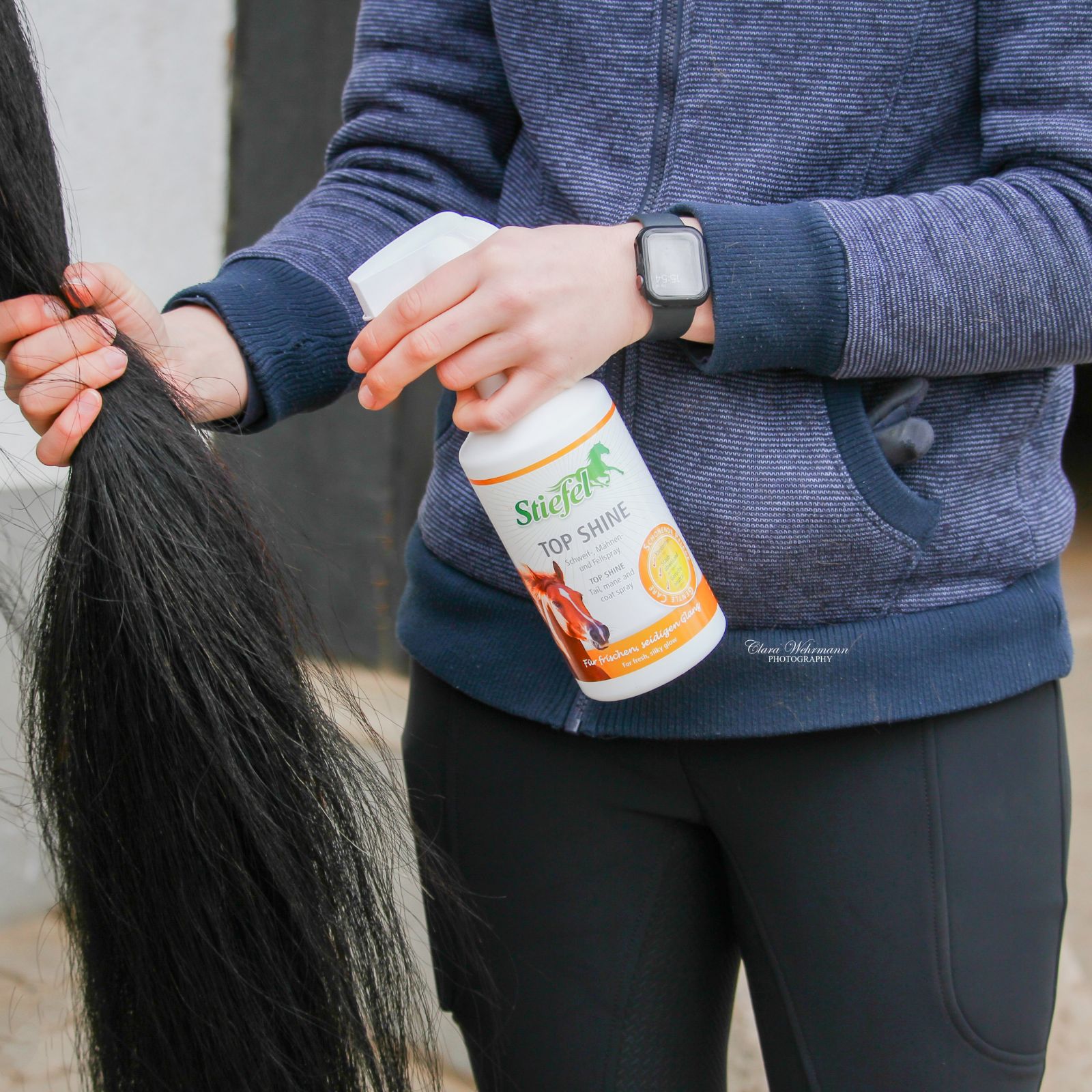 Person hält eine Sprühflasche TOP SHINE. Schwarzer Pferdeschwanz. Stiefel Logo.
