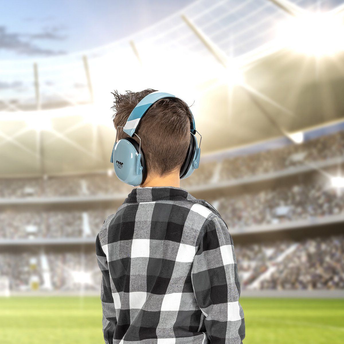 Bambino con cuffie blu davanti allo stadio. Indossa camicia a quadri. Cuffie con marchio reer.
