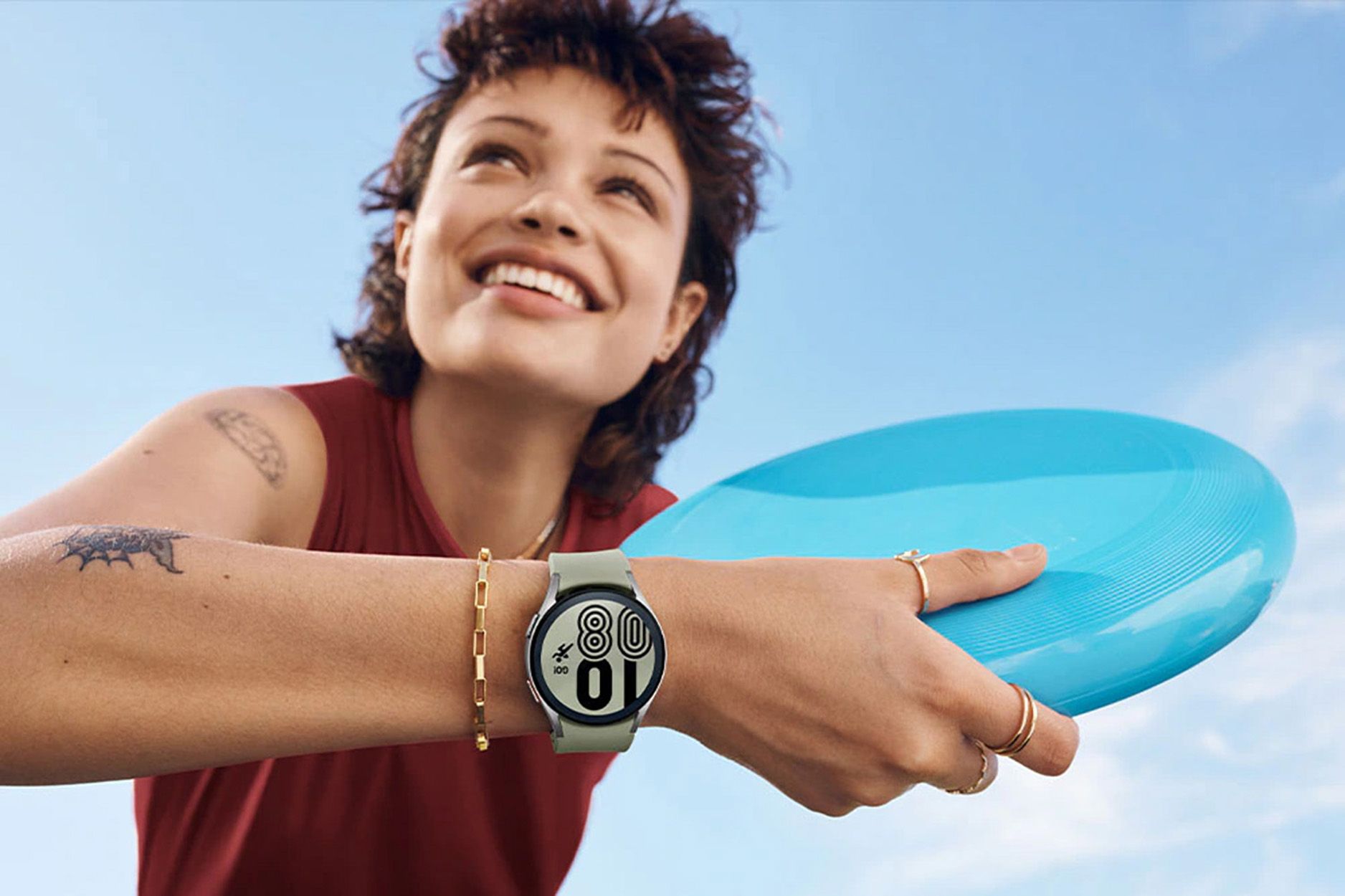 Frau mit Smartwatch am Handgelenk, hält Frisbee. Helle Smartwatch mit rundem Zifferblatt. Hintergrund: blauer Himmel.
