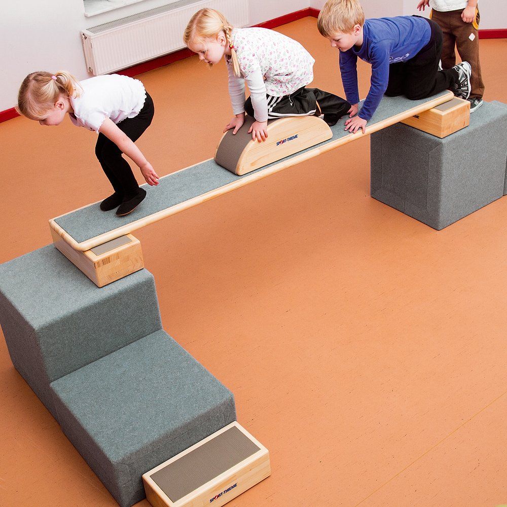 Kinder balancieren auf einer Holzkonstruktion. Mehrere Schaumstoffelemente bilden eine Treppe. Innenraum.